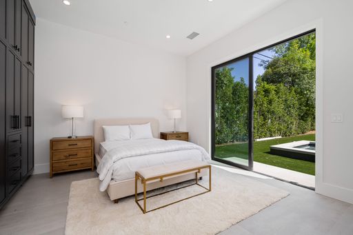 Modern bedroom with large glass doors opening to a backyard, featuring a comfortable bed, wooden furniture, and a neutral color palette.