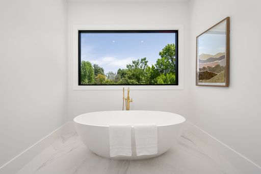 Modern bathroom with a white freestanding bathtub, large window with a nature view, and framed artwork on the wall.