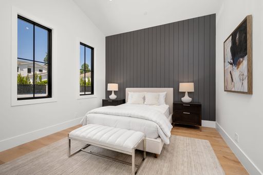 Modern bedroom interior with a gray accent wall, white bedding, and two windows showing a clear sky and neighborhood houses.