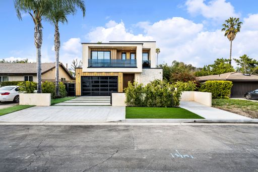 Modern two-story house with large windows, palm trees, and a clean driveway under a bright blue sky.