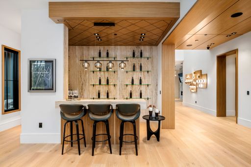 Modern home bar with wooden accents, bar stools, and glass shelves displaying wine bottles in a contemporary setting.