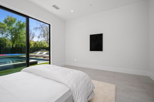 Modern minimalist bedroom with large window overlooking a pool and outdoor seating area, featuring white walls and simple decor.