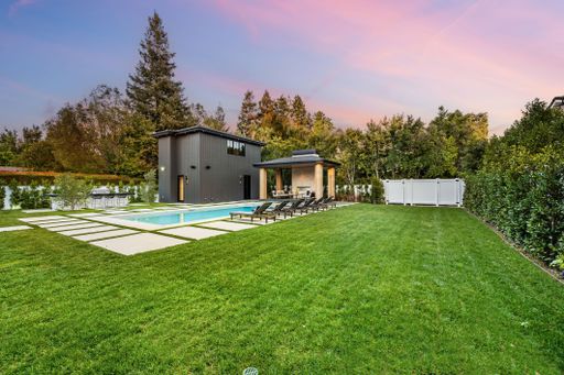 Modern backyard with a swimming pool, lounge chairs, and lush green lawn during sunset, surrounded by trees.