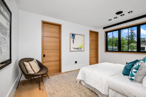 Modern bedroom interior with wooden doors, large window, and contemporary decor including a chair and abstract painting.