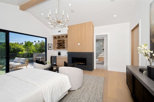 Modern bedroom with large windows, a balcony view of greenery, and contemporary decor including a chandelier, fireplace, and wooden accents.