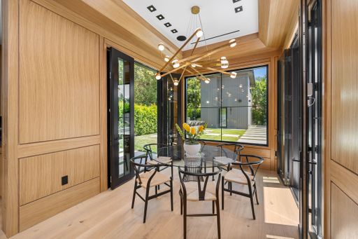 Modern dining area with wooden walls, glass table, black chairs, and a contemporary chandelier, overlooking a lush green garden.