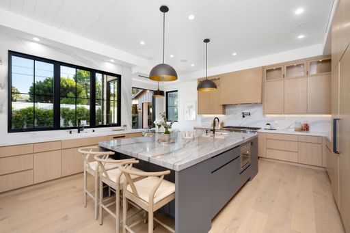 Modern kitchen with a large island, marble countertops, wooden cabinets, and large windows with black frames offering a view of greenery.