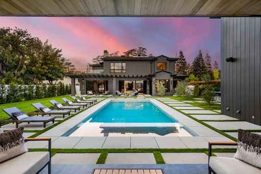 Modern luxury home with a large swimming pool, surrounded by lounge chairs and a lush garden, set against a vibrant sunset sky.