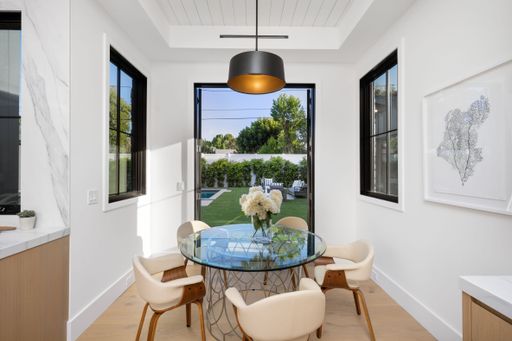 Modern dining area with glass table, stylish chairs, and large windows overlooking a backyard with greenery.