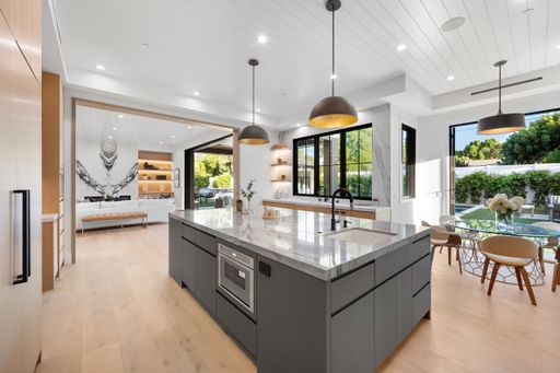 Modern open-concept kitchen and living area with large island, pendant lights, and floor-to-ceiling windows overlooking a lush backyard.