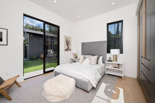Modern bedroom interior with large windows, neutral decor, and natural light, featuring a cozy bed, nightstands, and outdoor garden view.