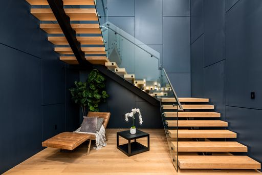 Modern interior with wooden floating staircase, glass railing, lounge chair, plant, and small table with flower arrangement.