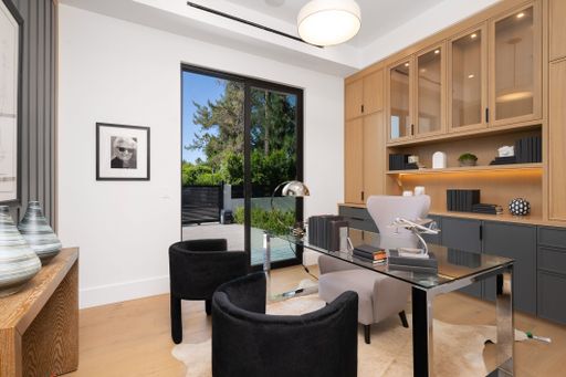 Modern home office with glass desk, black chairs, wooden cabinets, and large window overlooking green outdoor scenery.