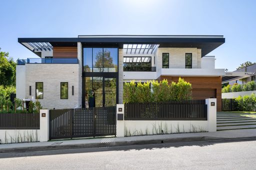 A modern two-story house with large windows, a black fence, and lush greenery in the front yard on a sunny day.