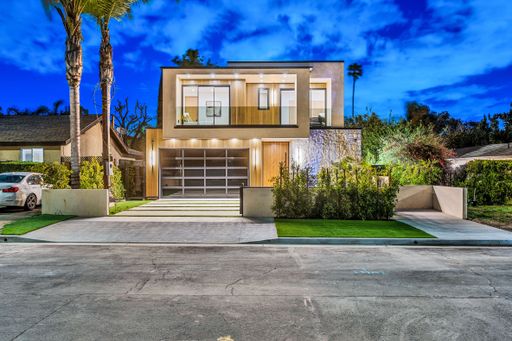 Modern two-story house with glass garage door and illuminated exterior at dusk, featuring palm trees and a clear blue sky.