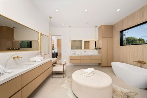 Luxurious modern bathroom with wooden cabinetry, marble countertops, a large mirror, and a freestanding bathtub, bathed in natural light.
