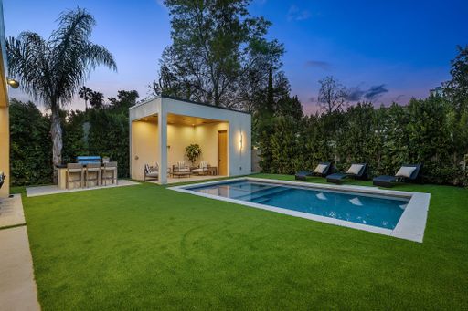 Modern backyard with a swimming pool, lounge chairs, and a covered patio area during twilight.