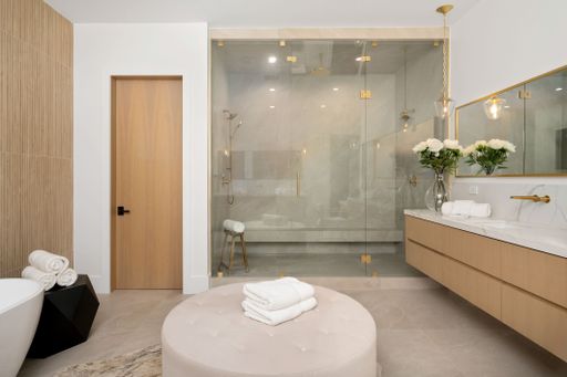 Modern bathroom interior with glass shower, wooden door, vanity with flowers, and a round ottoman with towels.