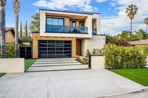 Modern two-story house with a glass garage door, lush greenery, and palm trees under a bright blue sky.