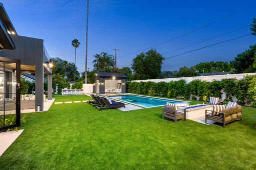 Modern backyard with a swimming pool, lounge chairs, and a cozy seating area, surrounded by lush greenery and palm trees.