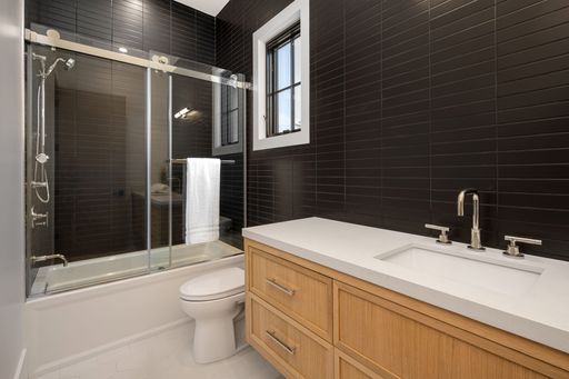 Modern bathroom with black tiles, glass shower enclosure, wooden vanity, and white countertop.