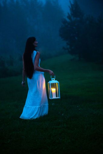 A woman in a white dress holding a lantern walks through a misty forest at dusk, creating a mystical atmosphere.
