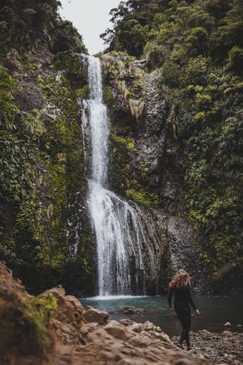 A vertical shot of a woman standing in front of the Kitekite Falls, New Zealand