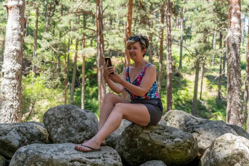 A Caucasian woman with short blonde hair looking at her phone screen while sitting on a rock
