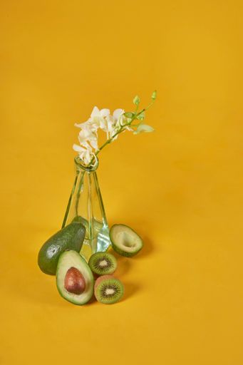 A vertical shot of a glass vase with flower, avocado, and kiwi on a yellow background