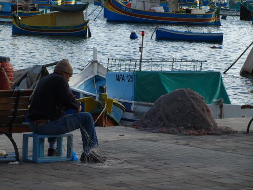A fisherman mending fishing net, along the coastal promenade in the fishing village of Marsaxlokk, Malta, on a cloudy winter day.