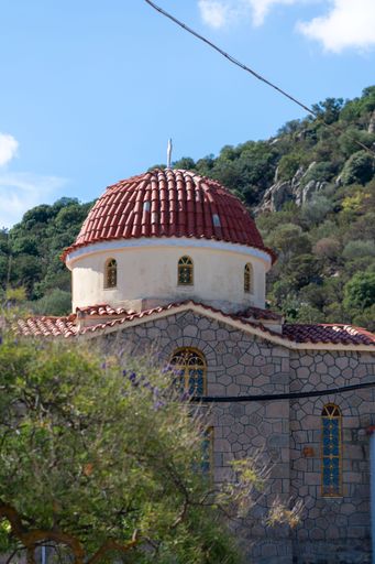 A traditional stone church with a red dome roof set against a backdrop of lush green hills under a clear blue sky