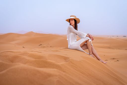 A woman in a white dress and straw hat sitting on a sand dune in a desert landscape under a clear blue sky.