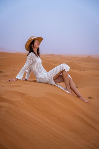 A woman in a white dress and straw hat sitting on a sand dune in the desert during sunset.