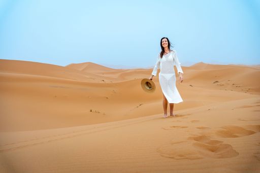 A woman in a white dress walking in the desert holding a hat with sand dunes in the background.