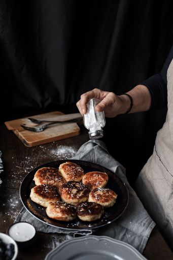 A vertical shot of a delicious pastry and a hand decorating it with powdered sugar arranged on a table with sour cream and blueberries