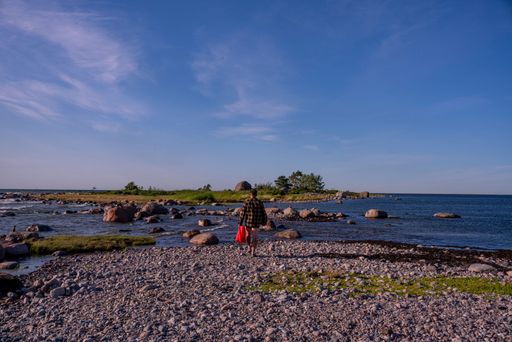 A person walking along a rocky shoreline under a clear blue sky, carrying a red bag, with distant greenery and calm sea.