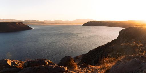 Panoramic view of a serene lake surrounded by rocky cliffs at sunset, with mountains in the background and a golden glow over the landscape.