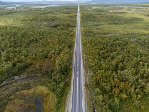 Aerial view of a long, straight road cutting through a lush green forest landscape, extending towards distant mountains under a cloudy sky.