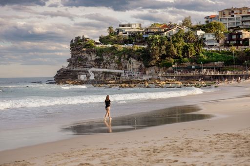 A woman walking along Bondi Beach with scenic cliffs and houses in the background under a cloudy sky.