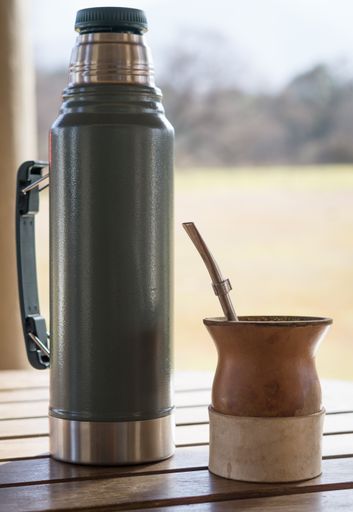 A vertical closeup of a cup of yerba mate infusion with a thermos.