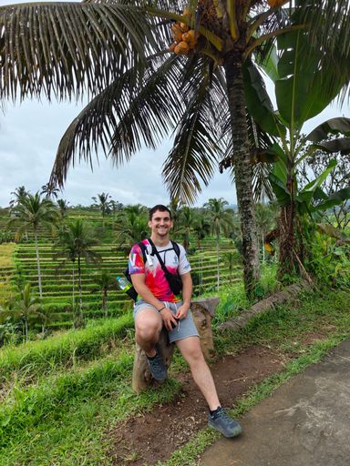 A young man sitting on a tree stump with lush greenery and fields behind.