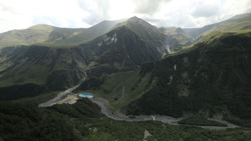 Scenic view of lush green mountains and a small lake under a partly cloudy sky.