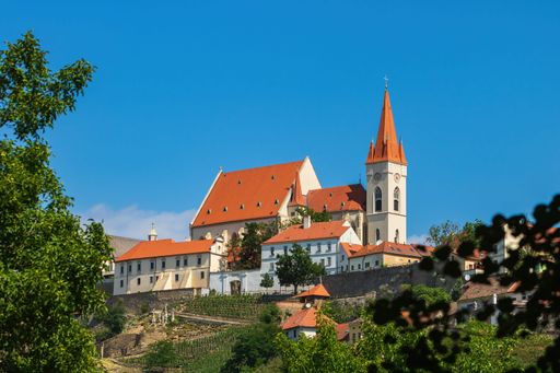 Scenic view of a historic church with red roofs surrounded by lush greenery under a clear blue sky in Znojmo, Czech Republic.