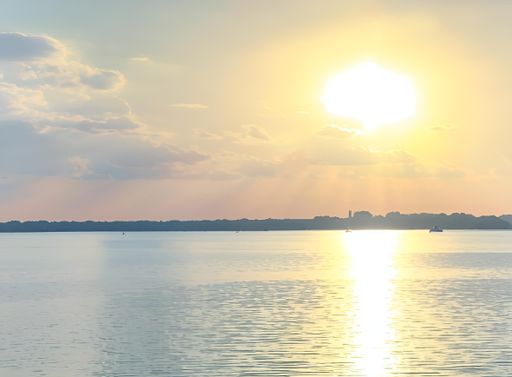 Serene sunset over a calm lake with boats and a distant shoreline, under a partly cloudy sky.