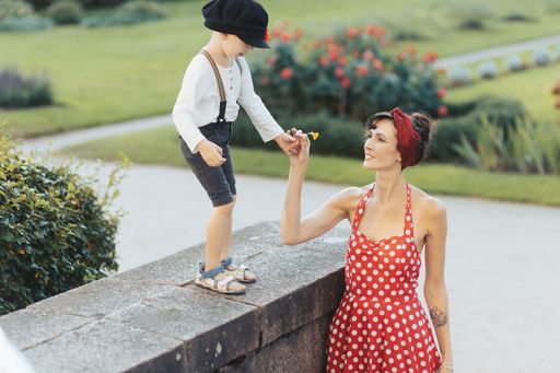 A young woman in a red polka dot dress receives a flower from a child in suspenders in a park.