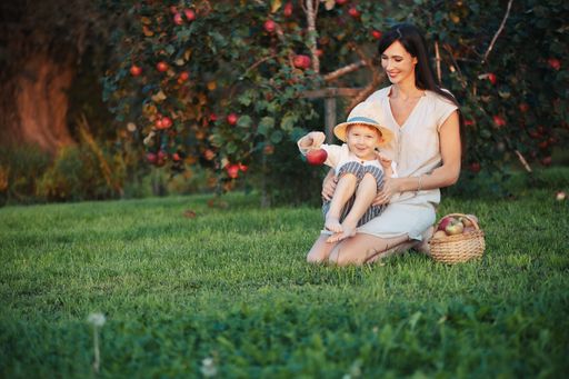 A mother and child enjoying apple picking in an orchard, with a basket of apples on the grass, during a sunny day.