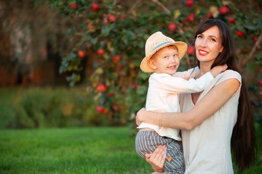 A young Caucasian mother holding her smiling son in a straw hat in an apple orchard on a sunny day.