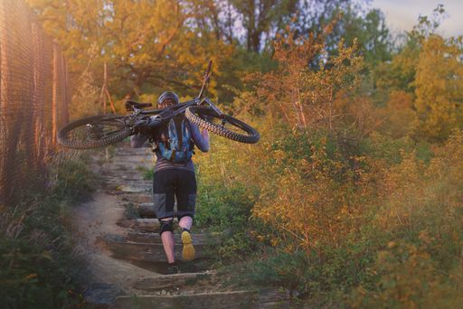 A cyclist carrying a mountain bike on his back uphill on wooden steps on a nature trail in the light of the setting sun.