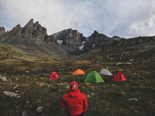 A person in a red jacket stands near colorful tents in a mountainous landscape under a cloudy sky.