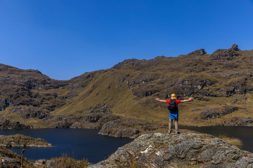 A hiker stands triumphantly on a rocky hill overlooking a serene lake and mountainous landscape under a clear blue sky.